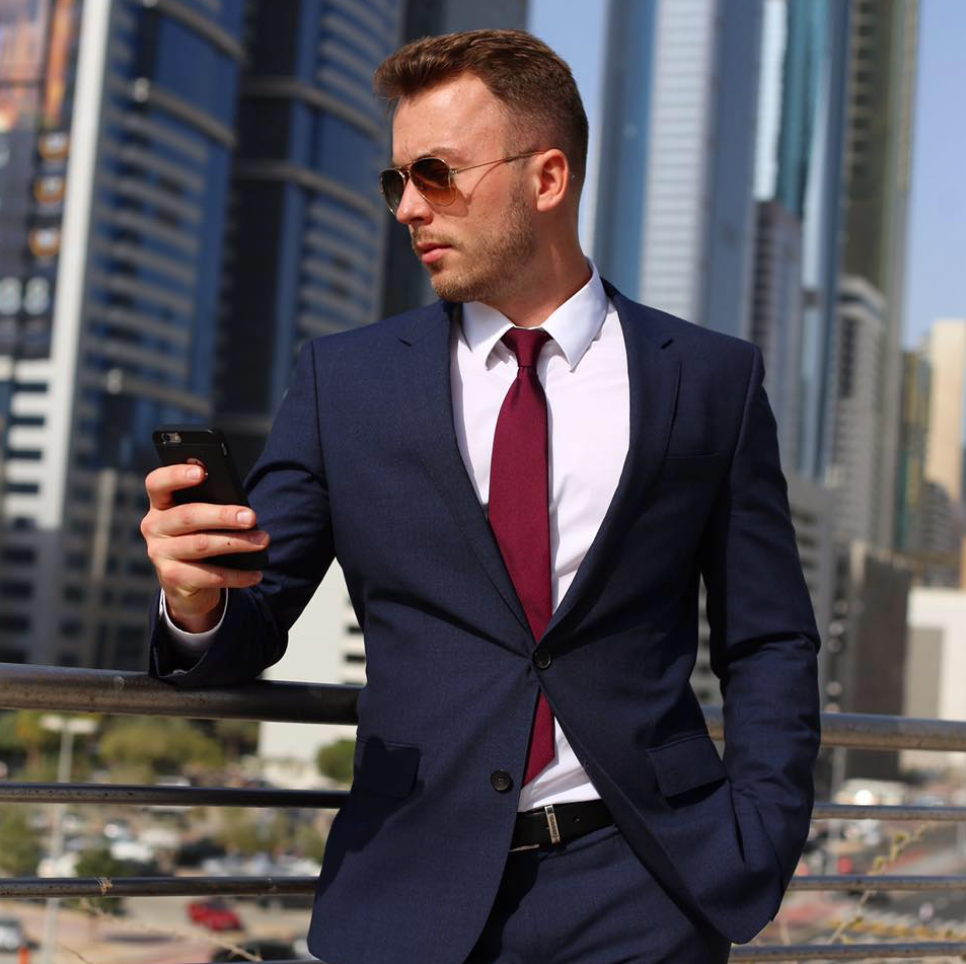 Максим Багрєєв в синьому класичному костюмі та червоній краватці стоїть на фоні сучасних будинків